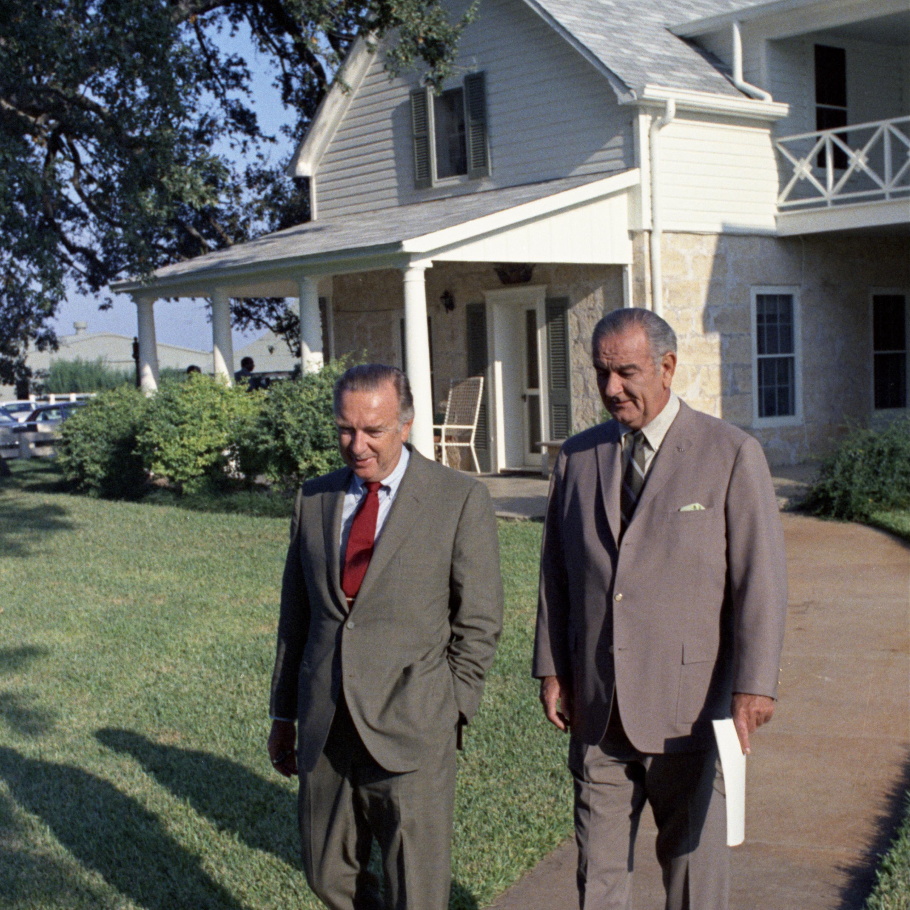 Walter Kronkite and Lyndon Baines Johnson walk down a sidewalk outside a country house. 